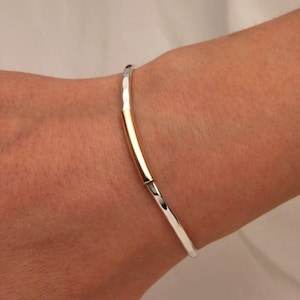 Może przedstawiać: Srebrna bransoletka typu bangle z złotym akcentem. Bransoletka jest otwarta i ma młotkowane wykończenie.