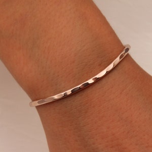 Pode incluir: Uma pulseira de bracelete martelada em ouro rosa. A pulseira tem uma superfície texturizada com pequenas reentrâncias irregulares. A pulseira curva parece ser de metal.