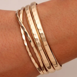 May include: A set of five gold-toned hammered metal bangles. The bangles are stacked on a person's wrist.