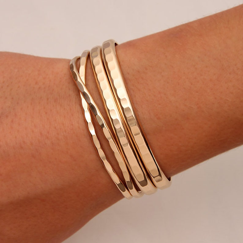 May include: A set of five gold-toned metal bangles, stacked on a person's wrist. The bangles are all different widths and have a hammered texture.