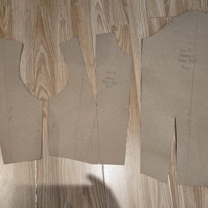Op de afbeelding: Drie bruine papieren naaipatronen zijn gerangschikt op een houten oppervlak. De stukken omvatten een lijfje voorkant, een lijfje achterkant en een mouwpatroon. Handgeschreven notities en markeringen zijn zichtbaar op de patronen.