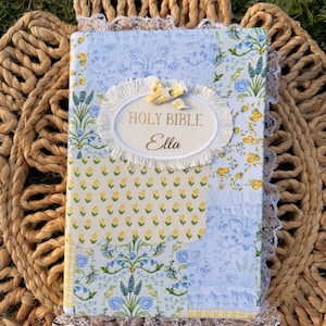 Puede incluir: Una Biblia con estampado floral en diseño azul claro y amarillo. La portada presenta el texto "Holy Bible Ella" en letras doradas, rodeado por un óvalo blanco con un ribete de encaje. Los bordes están adornados con encaje blanco.