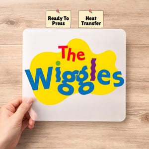 May include: A white heat transfer sheet with the words "The Wiggles" in blue, yellow, red, and purple. The words "Ready To Press" and "Heat Transfer" are printed on small labels above the sheet.