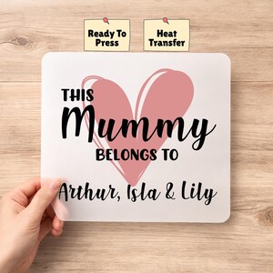 May include: A white square heat transfer featuring a pink heart and the text "THIS Mummy BELONGS TO Arthur, Isla & Lily" in black. Two small yellow labels read "Ready To Press" and "Heat Transfer".