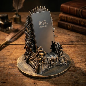 May include: A bronze-colored phone stand shaped like an iron throne, holding a smartphone displaying the time 8:15 PM, October 26. The stand is on a round base, with details of swords and spikes.