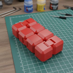 Pode incluir: Um brinquedo fidget vermelho composto por blocos interligados, repousando sobre uma base de corte verde. O brinquedo é projetado para ser flexível e manipulável. Frascos de tinta e ferramentas são visíveis no fundo.