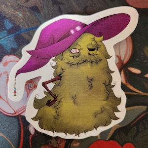 May include: A sticker featuring a whimsical, cartoonish creature with a yellow-green, furry body and a purple hat. The creature has one visible red eye and a thin, brown arm. The sticker is on a floral patterned surface.