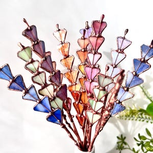 May include: A bouquet of colorful stained glass flowers in shades of blue, purple, orange, pink, and green. The glass pieces are arranged on copper stems, creating a unique and artistic floral arrangement. The flowers are displayed in a white vase.