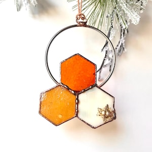 May include: A stained glass ornament shaped like a honeycomb with three cells. The top cell is orange, the left cell is yellow, and the right cell is white with a gold bee. The ornament is suspended from a copper wire loop.