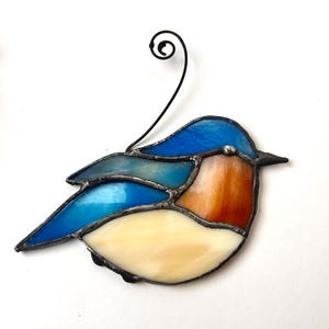 May include: Stained glass bird ornament featuring a blue head and wings, an orange breast, and a cream-colored belly. The bird has a black metal outline and a wire for hanging. A decorative piece for home decor.