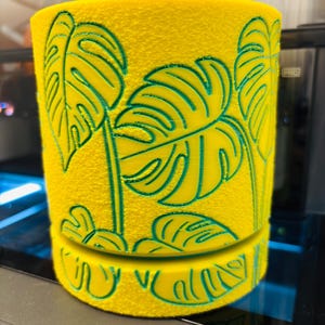 Pode incluir: Um vaso cilíndrico amarelo com uma superfície texturizada e padrões de folhas de monstera verdes em baixo-relevo. O design inclui uma costura horizontal perto da base, separando o corpo principal da parte inferior, com motivos de folhas que continuam por ambas as partes.