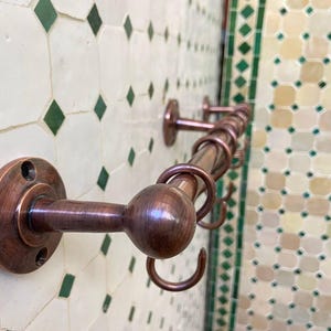 May include: A copper-colored towel rack with multiple hooks, mounted on a wall with a decorative tile pattern. The rack has a cylindrical bar and round hooks, providing a stylish storage solution for bathrooms or kitchens.