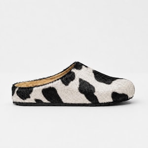 Puede incluir: Una zapatilla tipo mule con un patrón de estampado de vaca en blanco y negro en el exterior. El calzado tiene una punta redondeada, una silueta de perfil bajo y un forro interior de color beige, sobre una suela de goma negra texturizada.