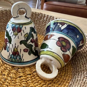 May include: Two ceramic bells with floral designs. One bell is upright, the other is on its side. The bells are white with blue, red, and green accents. They have a loop at the top for hanging. The bells are sitting in a woven basket.