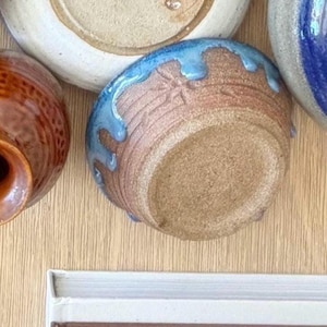 May include: A collection of handmade ceramic bowls in various sizes and colors. The central bowl is brown with a blue glazed rim. Other bowls include a white and tan bowl, and a blue and white bowl. The bowls are arranged on a light wood surface.