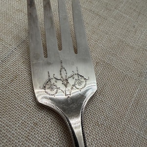 Puede incluir: Primer plano de un tenedor plateado con cuatro púas. El mango presenta un diseño grabado ornamentado, con motivos florales y de volutas. Las púas están ligeramente deslustradas, mostrando signos de edad y uso. El tenedor es probablemente antiguo.