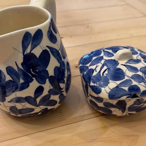 May include: A white ceramic pitcher and a covered dish with a blue floral pattern. The pitcher has a handle and a spout. The dish is oval-shaped with a small knob on top. Both pieces are on a light-colored wooden surface.