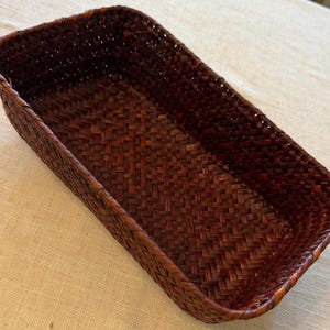 May include: A rectangular, woven basket in shades of brown. The basket has a tightly woven pattern and a slightly rounded rim. The basket is empty and sits on a light-colored surface. The basket is likely used for storage or display.