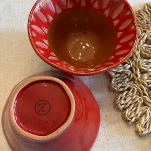 May include: Two red ceramic bowls. The top bowl has a red exterior with white leaf-shaped designs and a brown interior. The bottom bowl is red with a lighter rim and the text "MADE IN ITALY".
