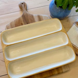 Puede incluir: Una fuente rectangular para servir con tres compartimentos, con interior color crema y exterior ámbar claro. La fuente descansa sobre una tabla de cortar de madera. Una pequeña tabla de cortar de madera y una maceta de cerámica azul con plantas verdes están en el fondo.