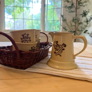 Puede incluir: Una jarra y cuencos de color crema con un diseño floral marrón están sobre una mesa de madera. Los cuencos están en una cesta de mimbre marrón. Una toalla a rayas blancas y beige está debajo de la jarra.