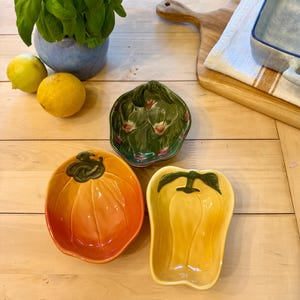 May include: A selection of ceramic vegetable-shaped serving dishes. Includes a pumpkin, artichoke, and bell pepper. The pumpkin dish is orange, the artichoke is green, and the bell pepper is yellow. Lemons and a basil plant are also present.