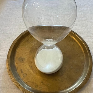 May include: A clear glass hourglass containing white sand, positioned centrally on a circular, weathered brass tray. The hourglass features a rounded top and bottom bulb connected by a narrow neck, with the white sand settled in the lower section.