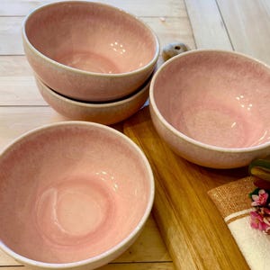 Puede incluir: Un juego de cuatro cuencos de cerámica rosa con acabado moteado. Los cuencos tienen forma redondeada e interior rosa claro. Los cuencos están apilados y colocados sobre una superficie de madera, con una tabla de cortar y una servilleta decorativa.