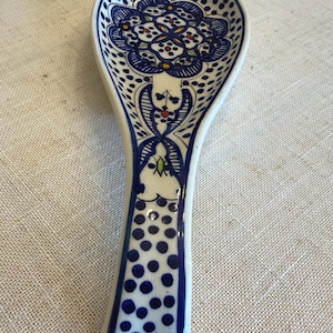 May include: A white ceramic spoon rest with a blue floral design. The spoon rest features a detailed pattern of blue dots and lines, with accents of red, yellow, and green. The handle is adorned with a series of blue dots.