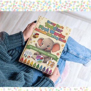 Puede incluir: Un libro para colorear para niños titulado "Buzzy Bee's Toddler First Words: Animals Coloring Book". La portada presenta una ilustración de un elefante de dibujos animados, una abeja y otros animales. El libro lo sostiene una persona con un suéter azul y jeans.
