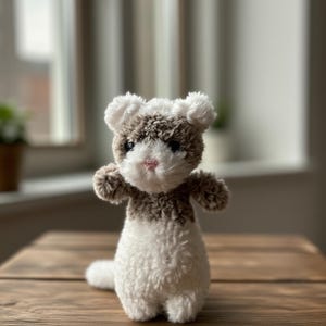 May include: A handmade crochet ferret plush toy. The ferret has a white body, a brown and white head, and small arms. The toy is sitting on a wooden surface. The ferret has a pink nose and black eyes.
