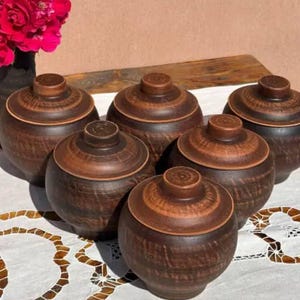 May include: Six handcrafted, brown ceramic jars with matching lids are arranged on a white lace tablecloth. The jars have a rounded shape and a textured surface. A vase with red flowers is in the background.
