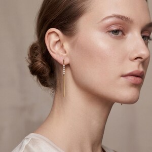 May include: A close-up of a woman wearing a gold earring. The earring features a vertical gold bar with a series of small, sparkling white gemstones. The earring is a simple, elegant design, suitable for everyday wear or special occasions.