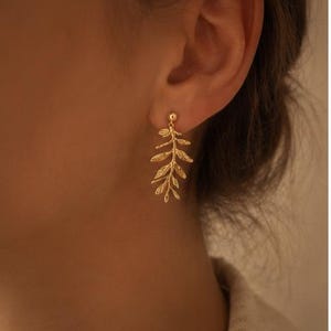 May include: A close-up of a gold-colored earring. The earring is a leaf design, with a small ball at the top where it connects to the ear. The leaf design hangs down from the ear. The background is a blurred image of a person's ear and shoulder.