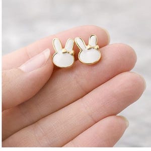 Może przedstawiać: Para złotych kolczyków sztyftów w kształcie królika. Każdy kolczyk ma białą emaliowaną twarz królika z małą kokardką. Kolczyki są trzymane w dłoni, pokazując ich rozmiar i design. Te kolczyki to uroczy i kapryśny dodatek.