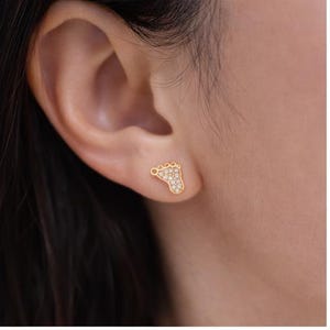 May include: A gold-colored stud earring in the shape of a baby foot, set with small, clear gemstones. The earring is worn on a person's ear, with the ear and part of the person's hair visible in the image.