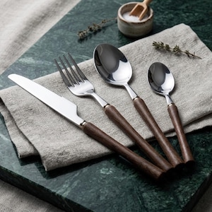 May include: A four-piece cutlery set comprising a serrated knife, a dinner fork, a large spoon, and a small spoon. Each item features a polished stainless steel head and a straight, dark brown wooden handle, arranged neatly on a folded linen cloth.