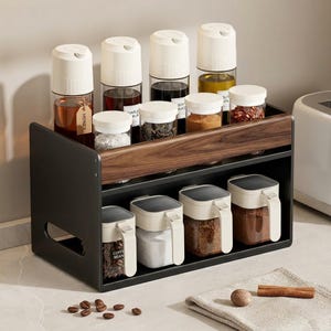 Puede incluir: Un especiero negro y de madera con botellas y frascos de vidrio transparente. La balda superior contiene botellas altas con tapas blancas y frascos más pequeños. La balda inferior tiene frascos cuadrados con asas. Algunos frascos contienen especias, aceites y granos de café.