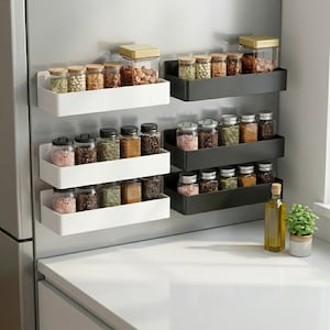 Könnte beinhalten: Eine Küchenszene mit magnetischen Gewürzregalen in Weiß und Schwarz, die an einem Kühlschrank befestigt sind. Die Regale enthalten verschiedene Gläser mit Gewürzen, Kräutern und Nüssen. Eine Flasche Olivenöl und eine kleine Topfpflanze stehen auf der Theke.