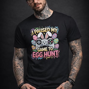 Könnte beinhalten: Schwarzes T-Shirt mit einer bunten Grafik, die einen Videospiel-Controller mit Hasenohren und Ostereiern zeigt. Der Text auf dem Shirt lautet "I PAUSED MY GAME TO EGG HUNT". Das Design ist im Cartoon-Stil gehalten.