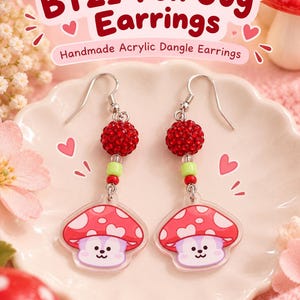 Könnte beinhalten: Hängeohrringe mit einem roten, pilzförmigen Acryl-Anhänger mit Cartoon-Gesicht, einer roten Strassperle und einer kleinen grünen Perle. Der Text BT21 Fun Guy Earrings Handmade Acrylic Dangle Earrings ist über dem Schmuckstück zu sehen.