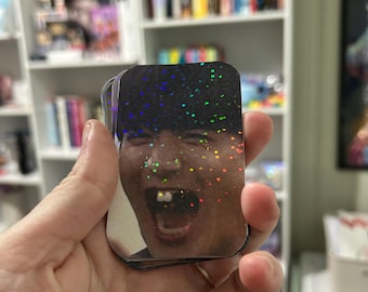 May include: A stack of rectangular stickers showing a person with an open mouth and missing teeth. The stickers have a holographic, glittery top layer with a dark background. A bookshelf is visible in the background.