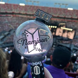 May include: A clear, spherical light stick with the name "Namjoon" and the BTS logo. The handle is black with a star pattern and the letters "BTS". The top has an "ARMY" label. The background shows a stadium full of people.