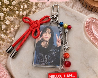 May include: A rectangular photo bag charm featuring a portrait of a person making a peace sign, a red cord bow, and a silver chain with colorful beads, a microphone charm, and a star. A second rectangular tag reads HELLO, I AM... KIM TAEHYUNG BIASED.