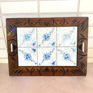 May include: A rectangular wooden serving tray with a carved border. The tray features six white tiles with a blue floral pattern. The tray has cut-out handles on each side. The wood has a dark brown finish.
