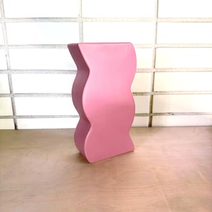 May include: A matte pink ceramic vase featuring a distinct wavy, undulating silhouette with a flat top and bottom. The object has a smooth, solid-colored surface and a vertical, serpentine shape that curves inward and outward along its sides.