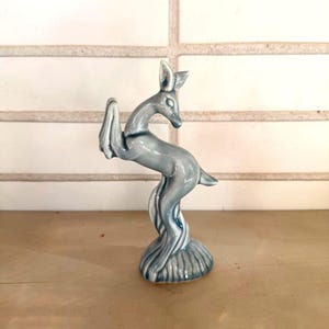 May include: A ceramic figurine of a deer in a leaping pose. The deer is a light blue-grey color with a glossy finish. The base is a textured, circular shape. The figurine is approximately 8 inches tall.