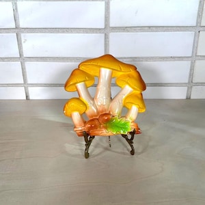 May include: A ceramic mushroom sculpture with yellow caps and white stems. The sculpture includes acorns and a green leaf, mounted on a decorative stand. The mushrooms are arranged in a cluster, creating a whimsical and naturalistic design.