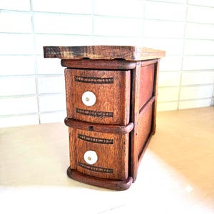 May include: A small, rectangular wooden storage unit featuring two stacked drawers. Each drawer front is crafted from dark brown wood with horizontal carved detailing and a single, round, white ceramic knob centered on the face.