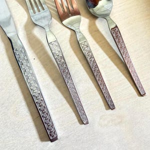 May include: A set of four stainless steel flatware pieces including a knife, two forks, and a spoon. Each handle features a repeating embossed floral pattern along the entire length. The metal has a polished finish with visible light reflections.